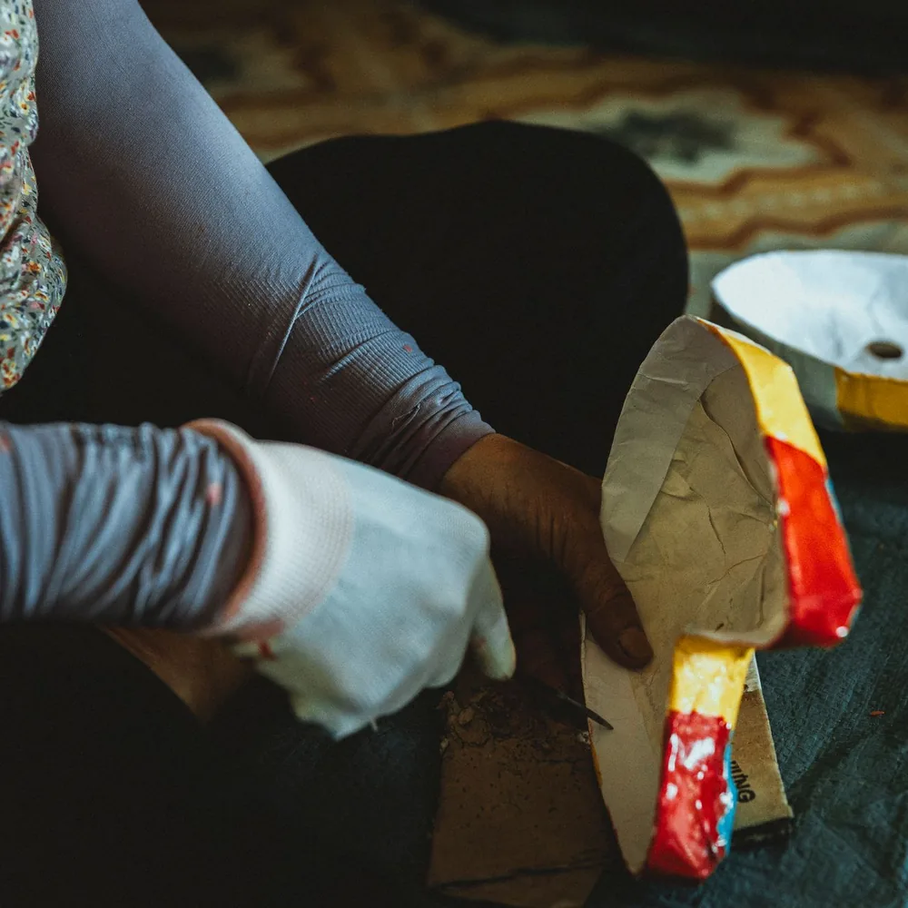 Manos trabajando una máscara de papel maché sobre una base recortada, con otra pieza secándose al lado
